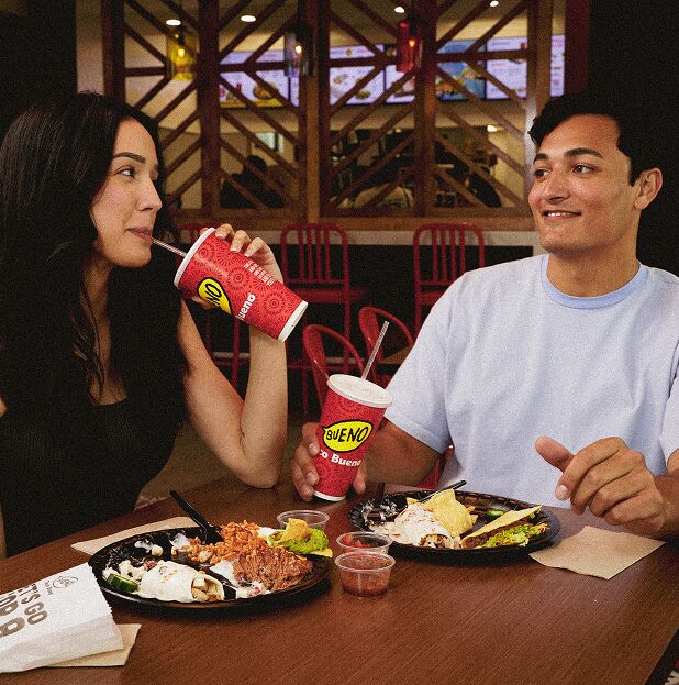 A young man and woman sharing a meal at Taco Bueno, enjoying tacos, burritos, and drinks while smiling at each other in a restaurant booth.