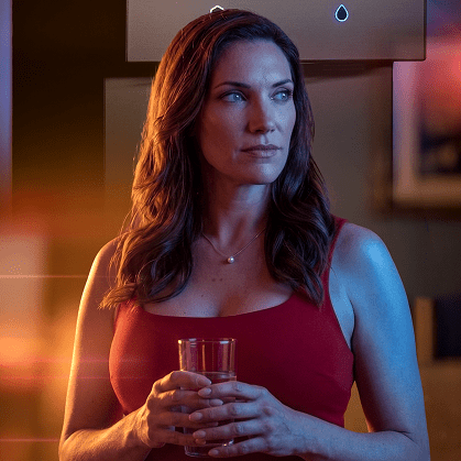 A woman with wavy brown hair wearing a red sleeveless top holds a glass of water, illuminated by warm orange and cool blue lights beside a modern water dispenser.