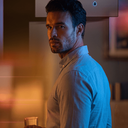 A man in a light blue button-down shirt turns to look over his shoulder with a serious expression, illuminated by dramatic warm orange and cool blue lighting inside a modern home.
