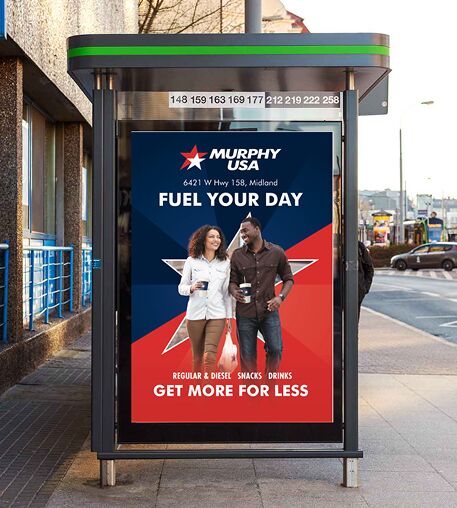 An outdoor advertisement for Murphy USA featured inside a bus shelter on a city street. The poster shows a smiling man and woman walking while holding drinks, under the text 'FUEL YOUR DAY' and near 'GET MORE FOR LESS' at the bottom.