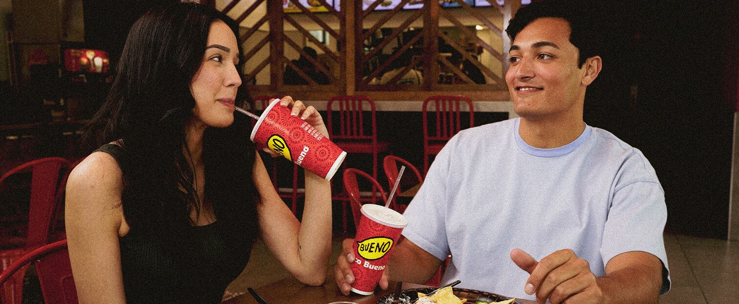 A young man and woman sitting at a table in a Taco Bueno restaurant, drinking from branded red cups and smiling.