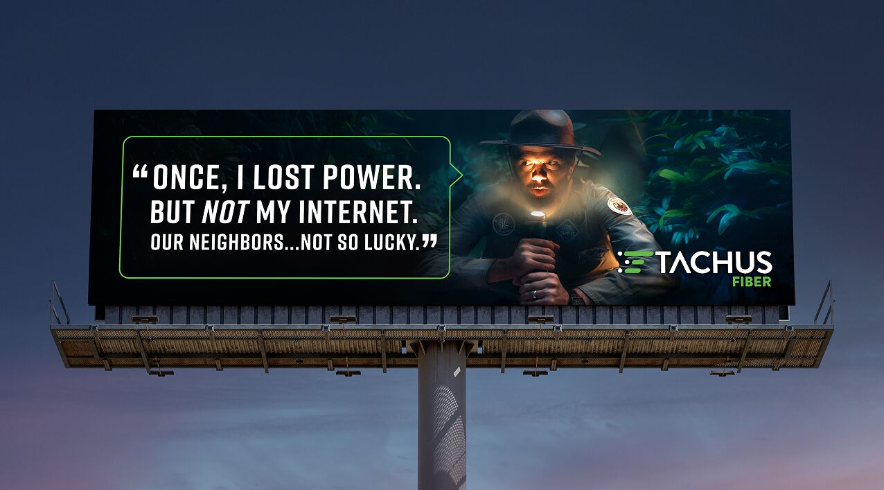 A billboard advertisement for Tachus Fiber featuring a man in a ranger hat shining a flashlight on his face in a dark, wooded setting. On the left, a speech bubble contains the quote, 'ONCE, I LOST POWER. BUT NOT MY INTERNET. OUR NEIGHBORS...NOT SO LUCKY.' The Tachus Fiber logo is displayed in the bottom right corner.