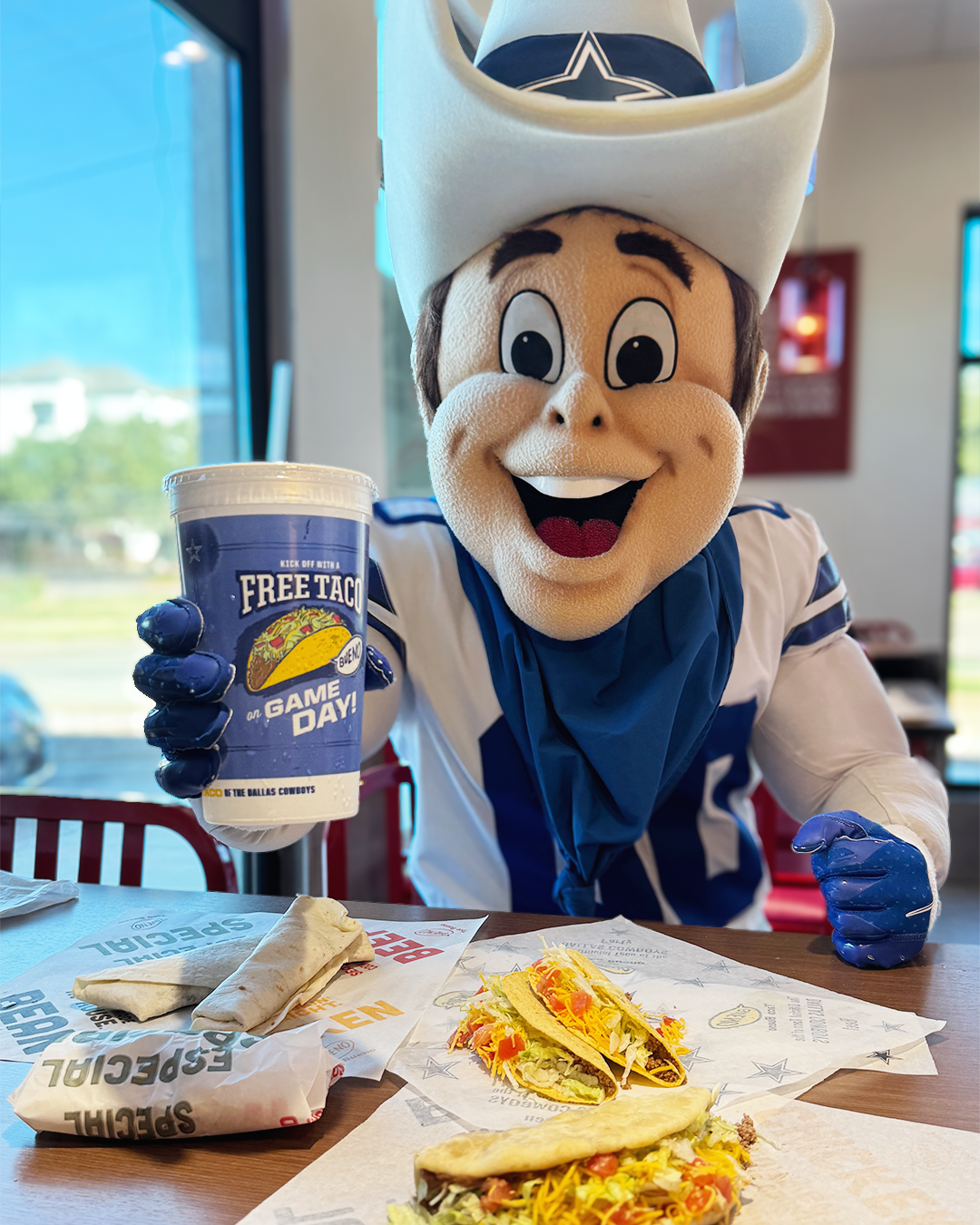 Dallas Cowboys mascot Rowdy smiling at a restaurant table, holding a blue cup promoting a "Free Taco on Game Day" next to spread out tacos and burritos.