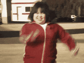 A smiling person with short dark hair jogs enthusiastically towards the camera wearing a red zip-up tracksuit jacket.