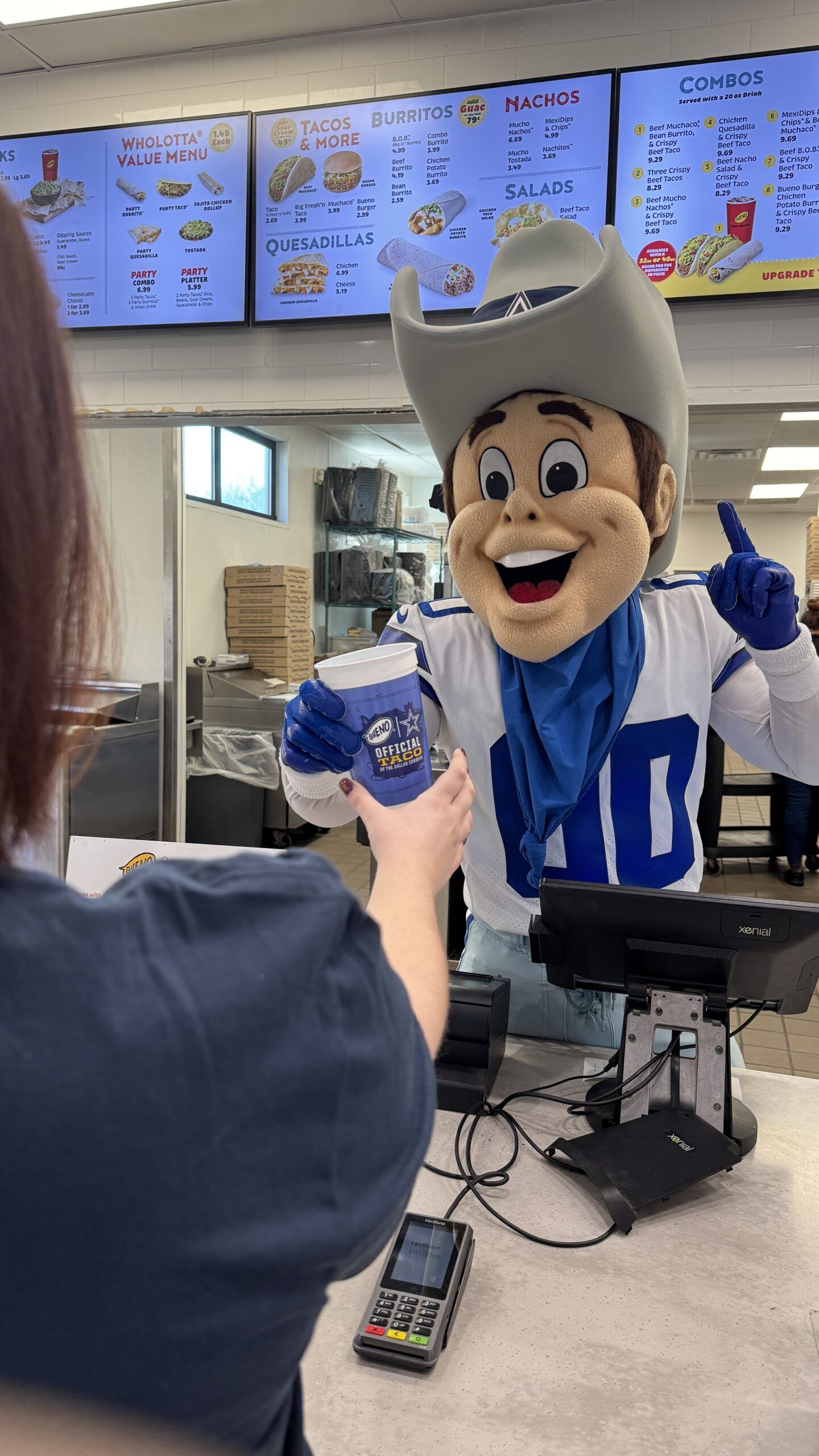 A smiling Dallas Cowboys-themed mascot wearing a cowboy hat, blue bandana, and white jersey hands a blue drink cup to a customer across the register counter at a Taco Bueno fast-food restaurant.