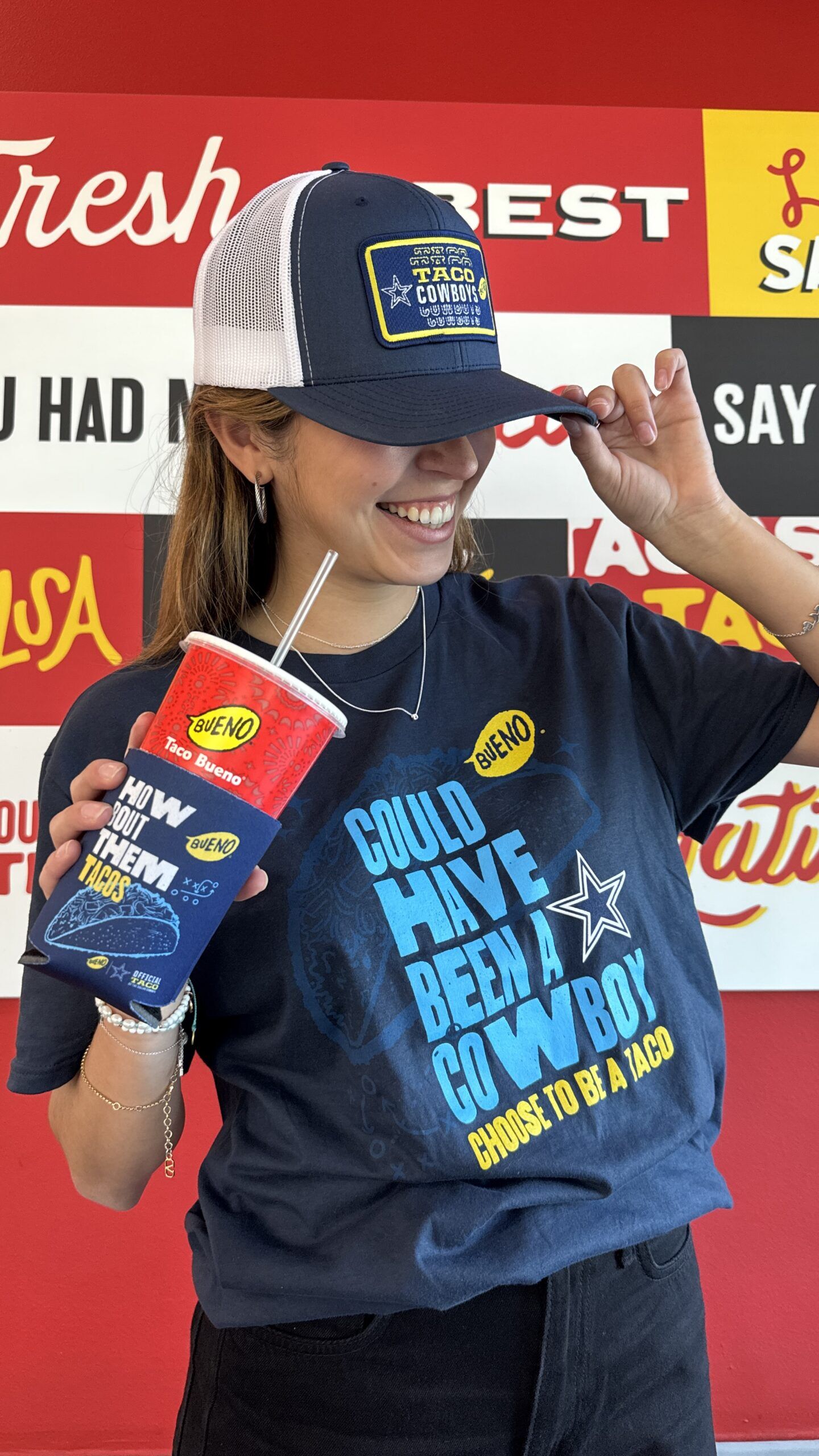 A smiling young woman wearing a navy blue 'Could Have Been A Cowboy, Choose To Be A Taco' t-shirt and matching trucker hat, holding a Taco Bueno drink cup with a 'How 'Bout Them Tacos' koozie.