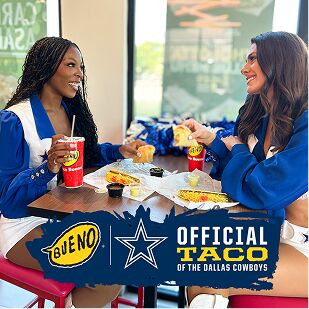 Two Dallas Cowboys Cheerleaders smiling and enjoying tacos at a table in a Taco Bueno restaurant, with a banner declaring Taco Bueno as the 'Official Taco of the Dallas Cowboys'.