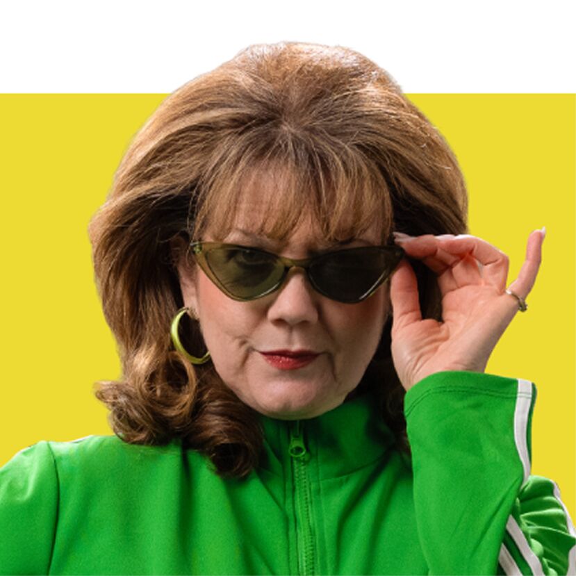 A woman with large, voluminous brown hair, wearing a green track jacket and stylish sunglasses, adjusts her sunglasses with her right hand while looking towards the camera. She has large hoop earrings and a ring on her right ring finger.