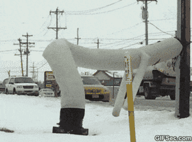 A white inflatable tube man repeatedly faceplanting and hitting its head against a wooden utility pole in a snowy parking lot.