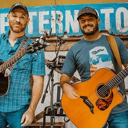 Two smiling male musicians posing with their acoustic guitars in front of a weathered blue sign.