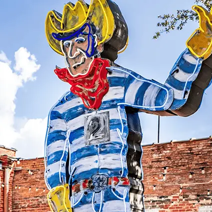 A vintage neon sign of a waving cowboy wearing a yellow hat, a red neck kerchief, and a blue-and-white striped shirt, set against a brick background and a blue sky.