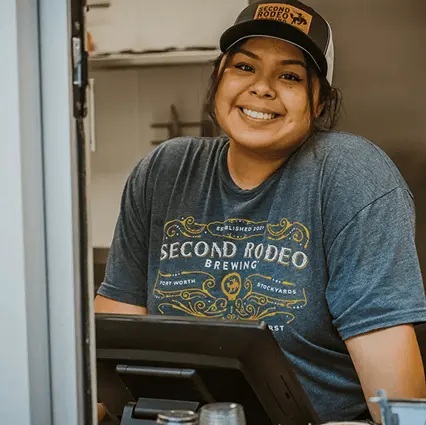 A friendly, smiling woman wearing a Second Rodeo Brewing t-shirt and trucker hat behind a service counter.