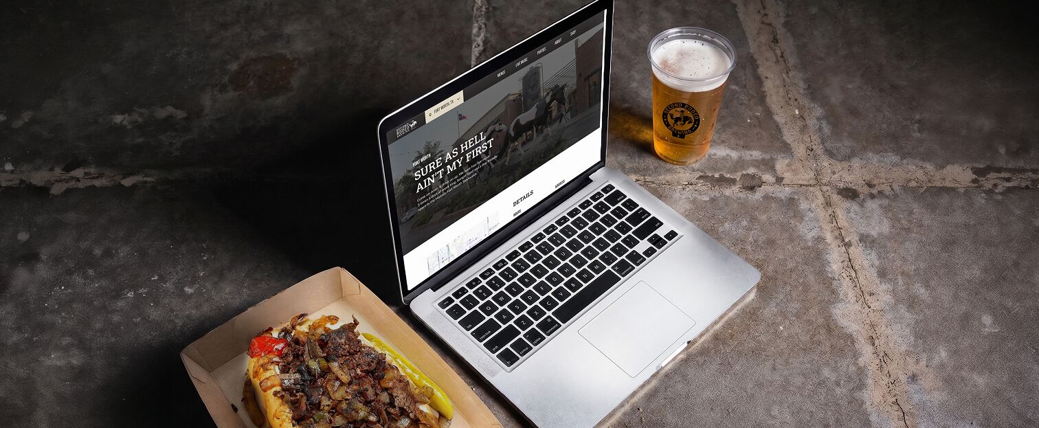 A silver laptop open to a website titled "Second Rodeo Brewing" with the tagline "Sure as hell ain't my first.", next to a delicious-looking cheesesteak and a pint of beer.
