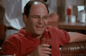 A man with glasses and a red shirt is laughing heartily with a drink in his hand.