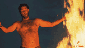 A shirtless, wild-eyed Tom Hanks from Cast Away standing triumphantly with arms outstretched next to a large, roaring bonfire at night.