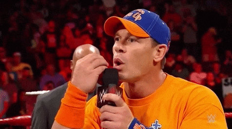 Close-up of John Cena in WWE arena, talking into a microphone, gesturing with his hand near his mouth.